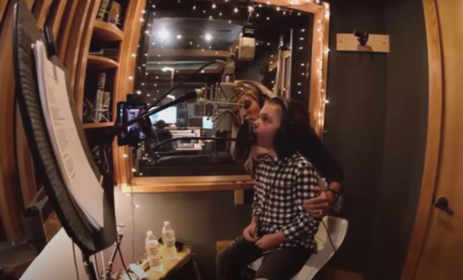 Inside a recording studio, we see Isaiah Fisher sitting in front of a mic singing. His mom, Carrie Underwood, stands behind him, hands on his arms in support.