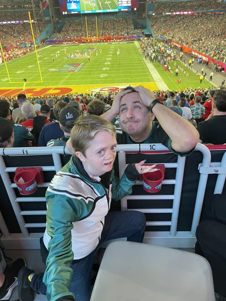 Kellirae Cox and Rick Santiago posing for a photo during their chance Super Bowl encounter. Rick has an over-the-top expression of stress on his face as he grasps his hair with his hands. Kellirae has a similar over-the-top look of annoyance, one hand out in a shrug motion. 