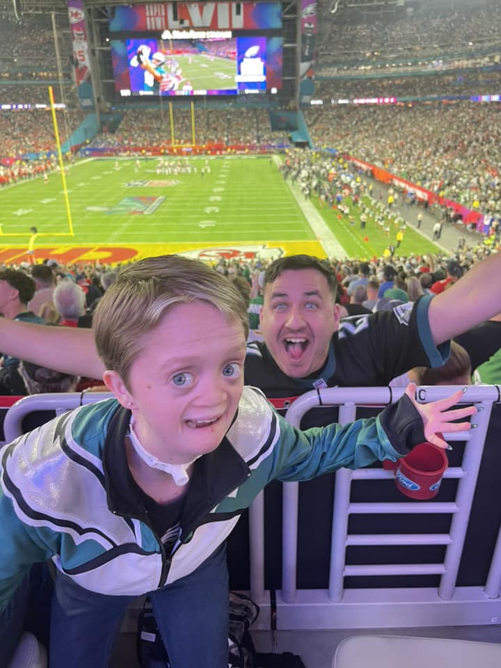 Kellirae and Rick smile, their arms stretched out wide, as they pose for a photo at the Super Bowl.