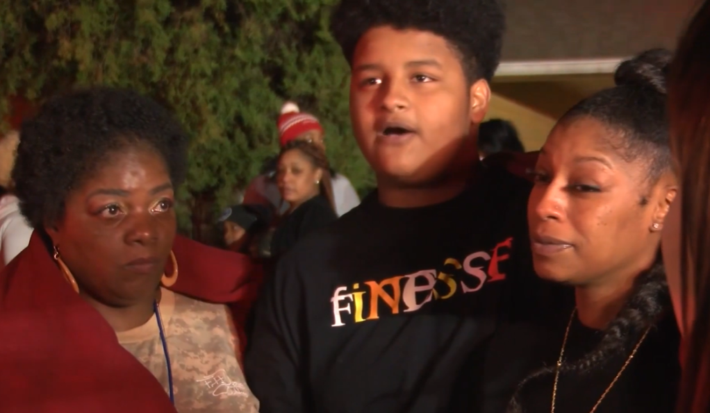 A woman, a teen boy, and a his mom stand side by side outside. The teen is talking.