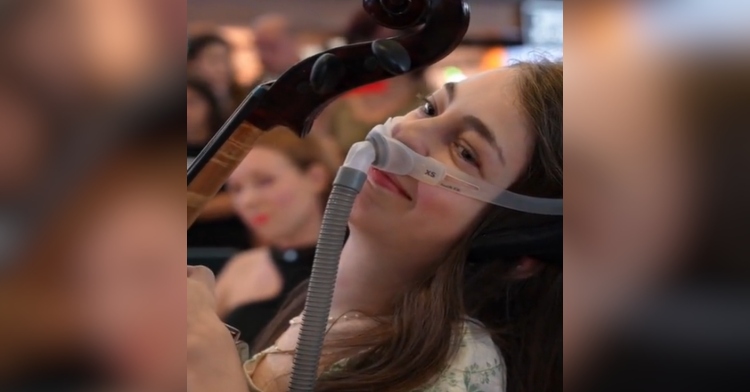 Close up of a disabled teen smiling as she plays the cello. There's a tube that's strapped around her face and goes into her nose.