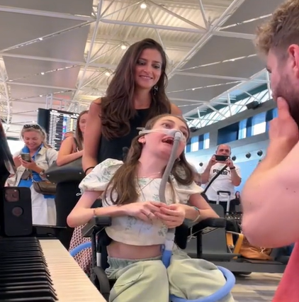 A disabled teen smiles as she talks to a pianist in the airport. The woman pushing her wheelchair smiles.