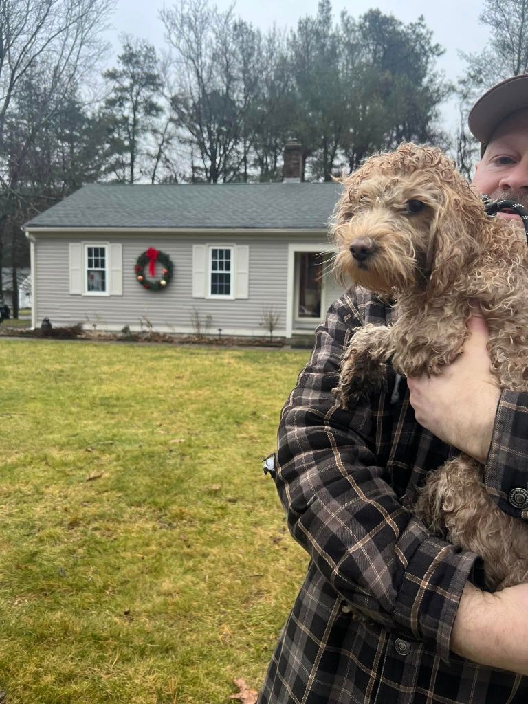 A man holds a dog, Margot. Her fur is curly and light brown in color. She looks dirty after her rescue. They're outside standing in front of a house. 