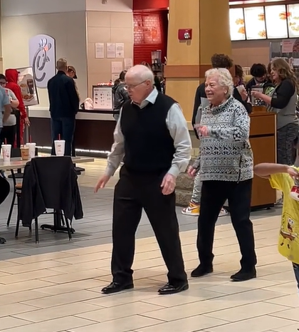 An elderly couple dance in the mall. The man is facing forward and his wife, who is dancing behind him, faces him. 
