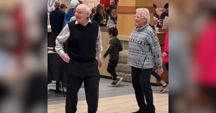 An elderly man looks up and over at something that can't be seen in the photo as he dances. His wife smiles as she dances toward him.