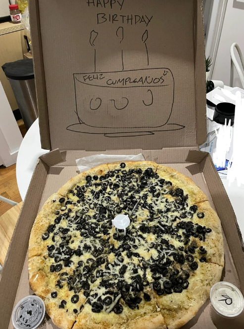 The pizza an employee made at the unique request of a customer that saved their birthday. The pizza is absolutely covered in black olives. To the side is a small container with more black olives. Inside the lid of the pizza is a drawing of a cake with "happy birthday" in English and Spanish.