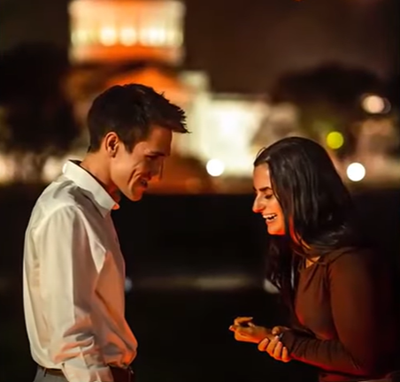 Bailey Davis leans forward, hand grasping her wrist as she smiles with her eyes closed while getting engaged. Her fiancé stands in front of her and smiles.