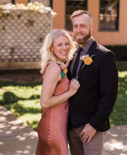 A woman, Brooke, and man, Johnathan, smile as they pose together outside. She's wearing a nice dress and he's in a suit.