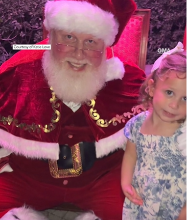 Steve Lantz, dressed as Santa, smiles as he poses for a photo with toddler Adley Love, who is also smiling.
