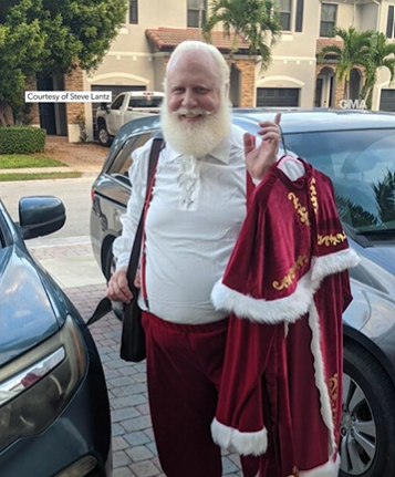 Steve Lantz, dressed as Santa, smiles as he stands next to cars outside. He's holding a hanger with an outer layer of his costume. 