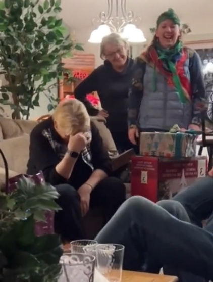 Grandma rests her face in her hands, head down, as she reacts to the Christmas prank her grandkids pulled off. Two women stand next to her, laughing. 