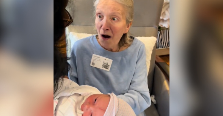 Grandma with dementia looks up at someone holding her newborn grandson. Grandma's eyes are wide and her mouth is agape.