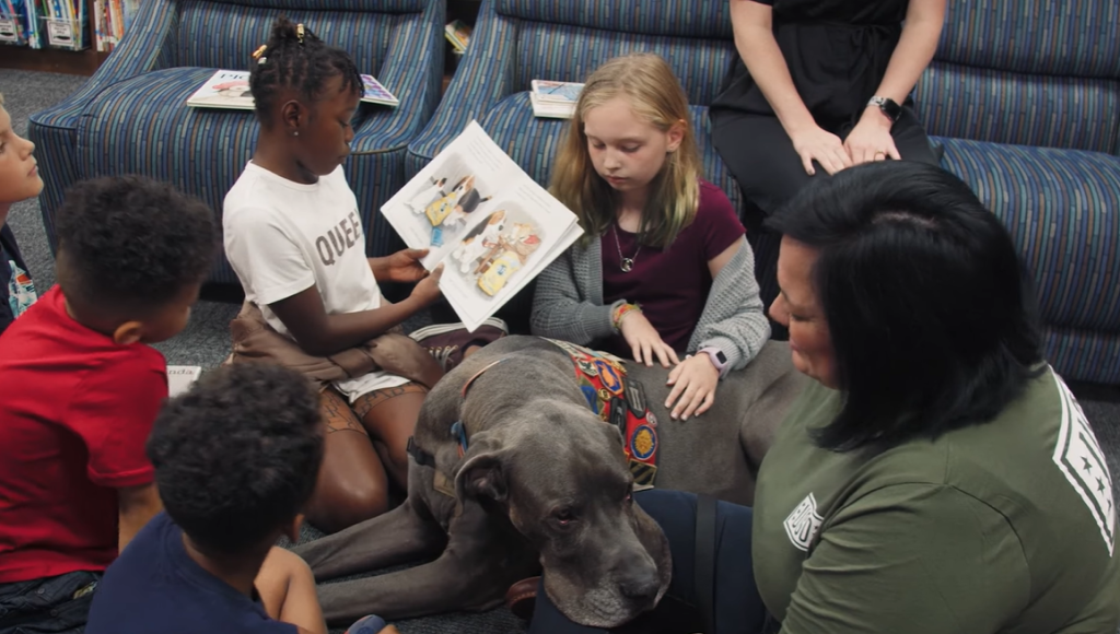 Maverick, hero dog of the year, lays down and rests his lap on a woman's knee amidst five children who sit next to him on the floor. One of the kids is reading from a book.