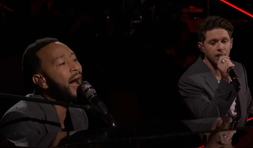 John Legend sits at a piano and sings into a mic as Niall Horan stands next to him, singing as well.