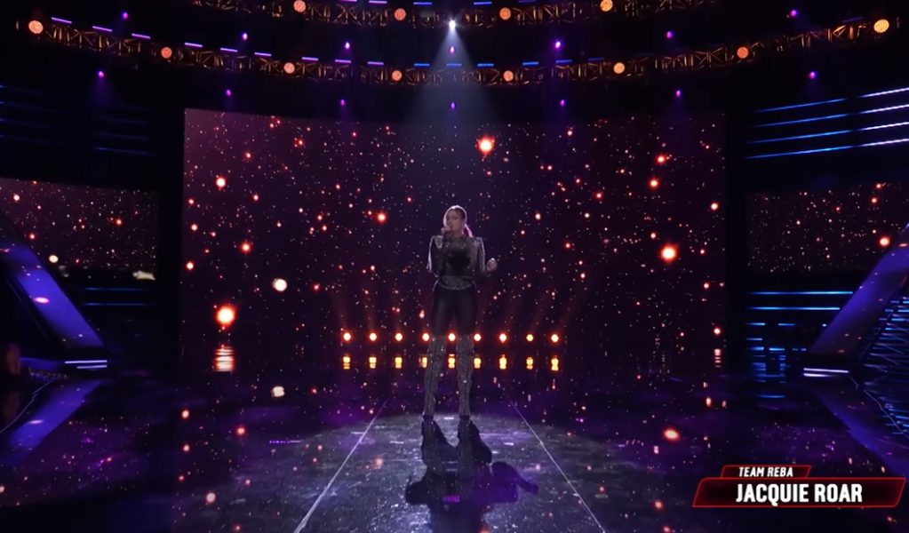Jacquie Roar sings on "The Voice" stage, sparkling lights swirling around her on the screens in the background.