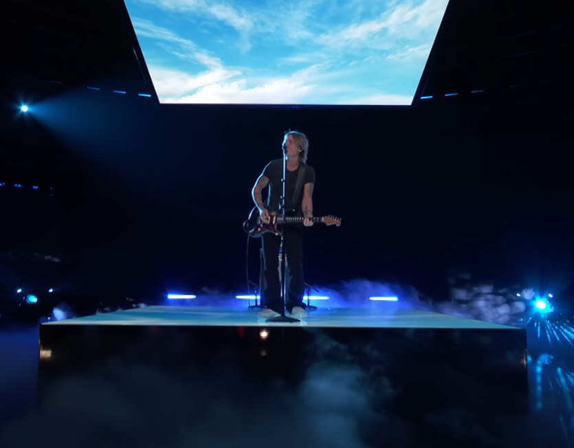 Keith Urban stands on an elevated, square stage with a screen above him that displays a blue sky with wispy clouds. He's performing "Blue Ain't Your Color" on the U.S. version of "The Voice."