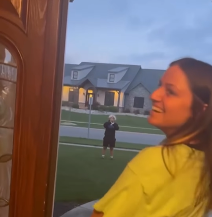 A teenage girl smiles as she looks back at the camera. She's at her front door, which is open. Standing at a distance in her front yard is a teenage boy playing a trumpet. 