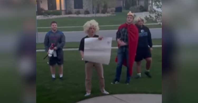 A teen named Gavin stands in a teen girl's yard with a large scroll. His three friends stand beside him. They're going for a "Monty Python" themed promposal.