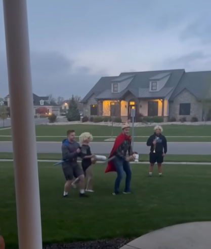 Four teenage boys are in a teen girl's front yard for a Monty Python themed promposal. One stands while holding a trumpet. The other three gallop in line, two acting as the front and end of a horse and the third acting like he's riding the horse.