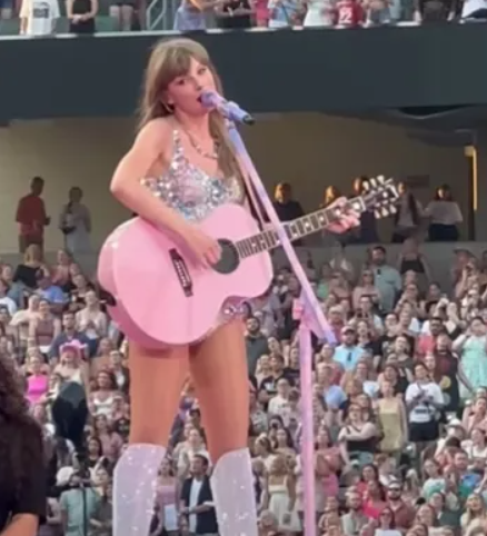 Taylor Swift sings into a mic as she plays a pink guitar during the Eras Tour.