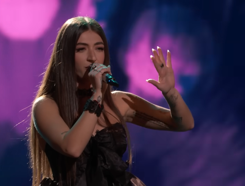 Nini Iris sings into a mic on "The Voice," one hand in the air out in front of her.
