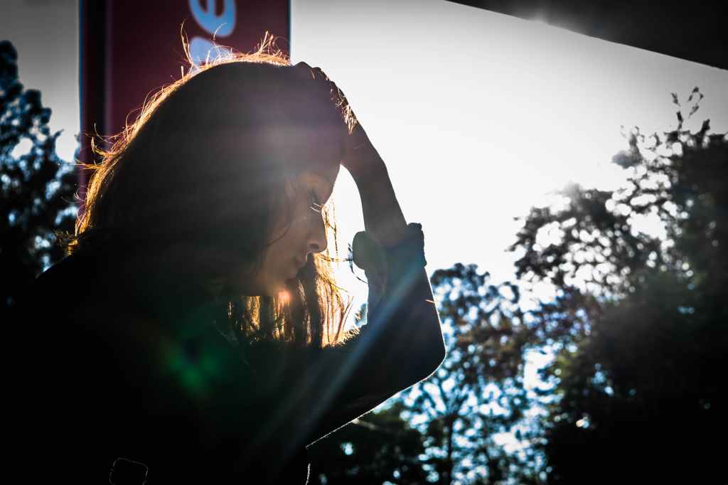 A woman stands near a window, looking down. A shadow is cast on much of her body and face. She looks sad as she pushes back her hair with one hand. Trees can be seen in the distance.