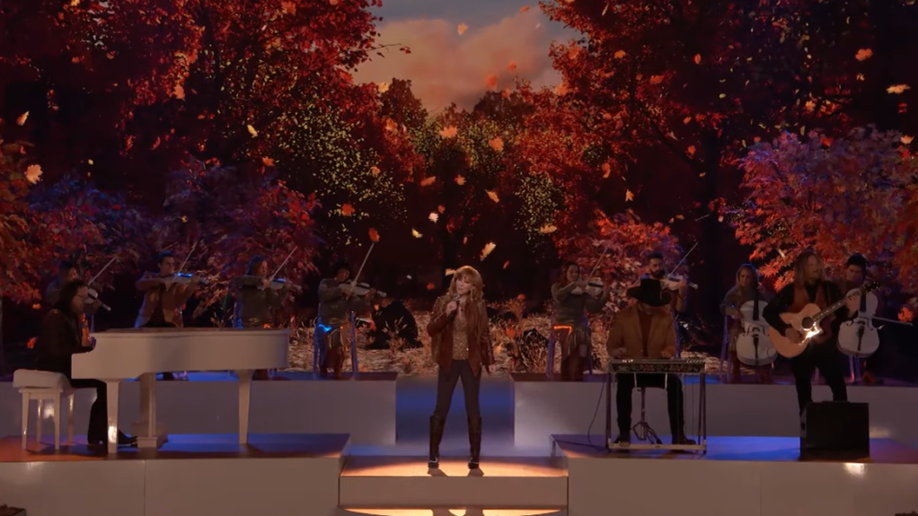 View of Reba on stage, singing her new song on "The Voice." Around her are folks playing instruments and, on screen, leaves fall. 