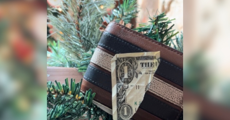 Close up of a corner of a dollar bill that has been ripped off. The bill is placed on a wallet which is placed among Christmas tree branches.