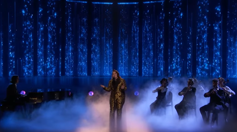 View from a distance of Ruby Leigh singing "Desperado" by the Eagles on "The Voice." Fog rises from the floor and a pianist and a group of violinists are on either side of the singer. On the screens behind her are streams of moving, blue lights.