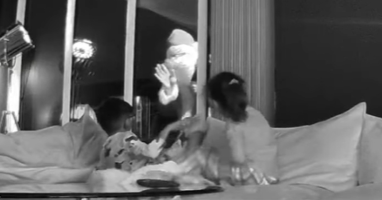 Two little kids sit on a couch and look out at Santa who is waving at them from the backyard. The image is black and white and comes from a Ring camera.