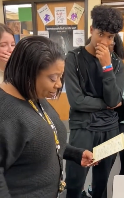 A teacher called Ms. Pow holds a card in her hand. Two of her students can be seen next to her, watching. Both have a hand over their mouth. One looks nervous and the other looks like they're getting emotional. 