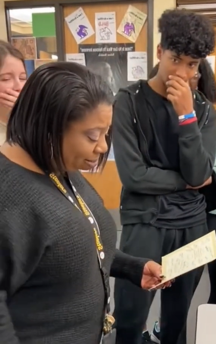 A teacher called Ms. Pow holds a card in her hand. Two of her students can be seen next to her, watching. Both have a hand over their mouth. One looks nervous and the other looks like they're getting emotional.