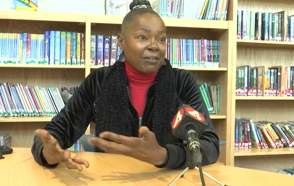 Teacher who saved a choking student, Tikiya Stewart, sits at a table in the school library and talks into a news mic.