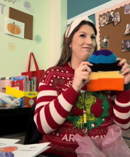 Teacher called Mrs. White smiles as she holds up a purple, blue, black, yellow, and orange striped hat. She's sitting at her desk at school and is wearing a festive Grinch sweater.
