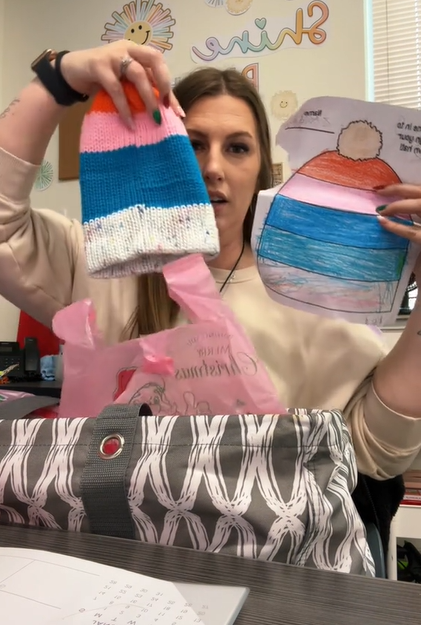 Teacher Mrs. White sits at her desk. In one hand she holds a hat that is orange, pink, blue, and at the bottom, white with colorful specks. In the other hand she holds a drawing of a hat that has been colored in and looks identical to the real hat she's holding.