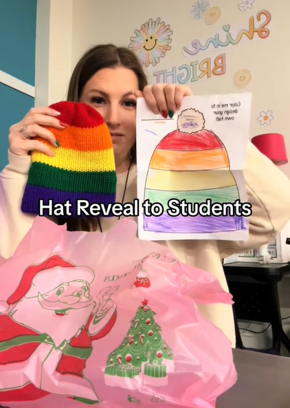 Mrs. White sits at her desk. With one hand she holds up a drawing a kid has colored in of a hat. In the other hand is a real-life version of the hat. The colors are red, orange, yellow, green, and purple.