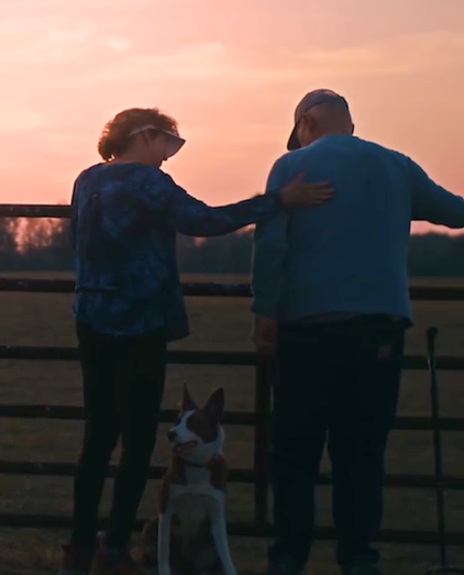 View from behind of Donald Adams, his wife, and Skippy standing on their farm at a fence. Donald's wife has a hand placed on his back. Skippy sits between them, facing the camera.