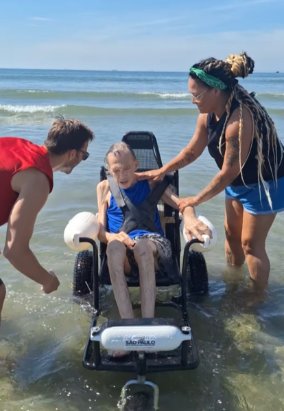 elderly man sits in a special wheelchair in shallow water on the beach as volunteers help him