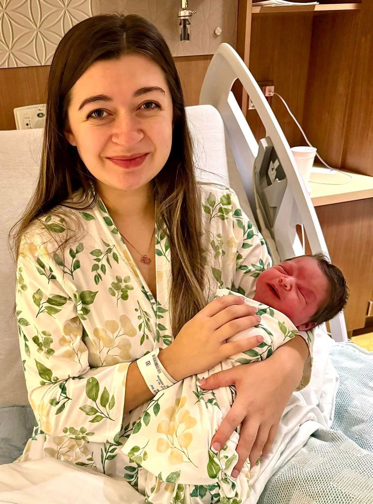 Analysia Beck smiles while sitting in a hospital bed. She's holding her baby, Micah, in her arms. The baby's eyes are closed and he's smiling a bit.