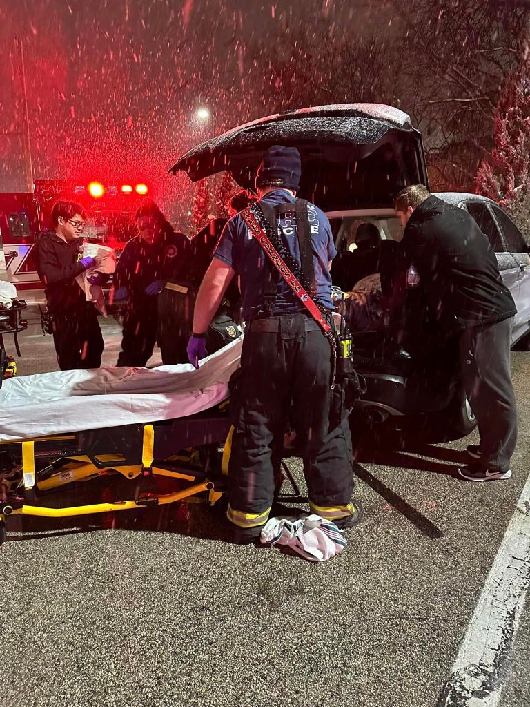 View of paramedics and other first responders gathered around the open trunk of Analysia and Daniel. A stretcher is being brough to the trunk and it's snowing.