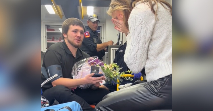 A man and a woman are sitting in the back of an ambulance. The man is holding back tears and has a bouquet of flowers as well as a box with an engagement ring. The woman is emotional, covering her face with her hands.