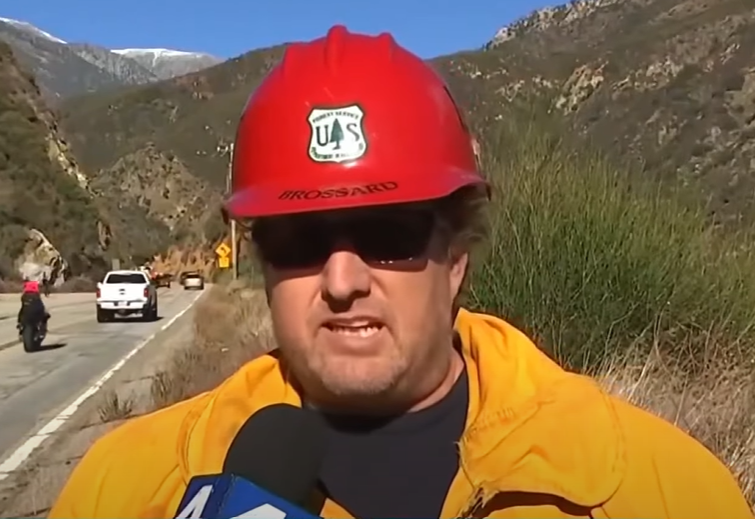 Matt Brossard, Engine Capt. of the U.S. Forest Service, wears a red work hat as he stands on the side of the road, mountains in the distance, as he talks into a mic about a cliff rescue.