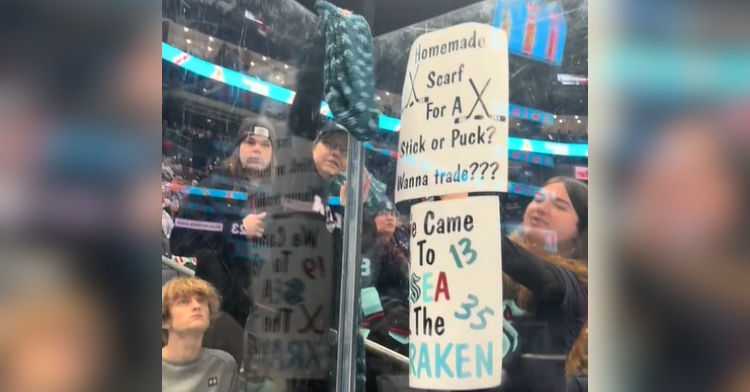 View of fans behind a clear barrier at a Seattle Kraken's game. One fan is holding up a sign that reads: "Homemade scarf for a stick or puck? Wanna trade???"