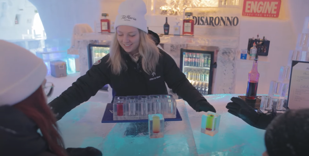 Ice Bar in an ice hotel called Hôtel de Glace. A woman smiles as she hands out colorful drinks in drinking glasses made of ice.
