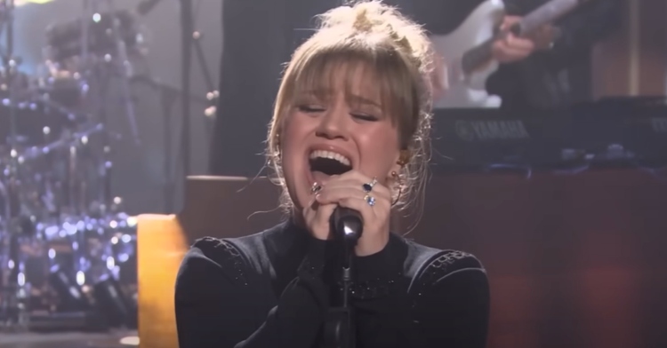 Close up of Kelly Clarkson singing passionately into a mic, her mouth wide open and her eyes closed.