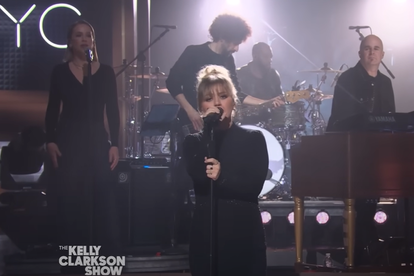Kelly Clarkson gazes into the camera with a serious look as she sings into the mic during a Kellyoke segment on her show. Folks playing instruments and singing background vocals are behind her.