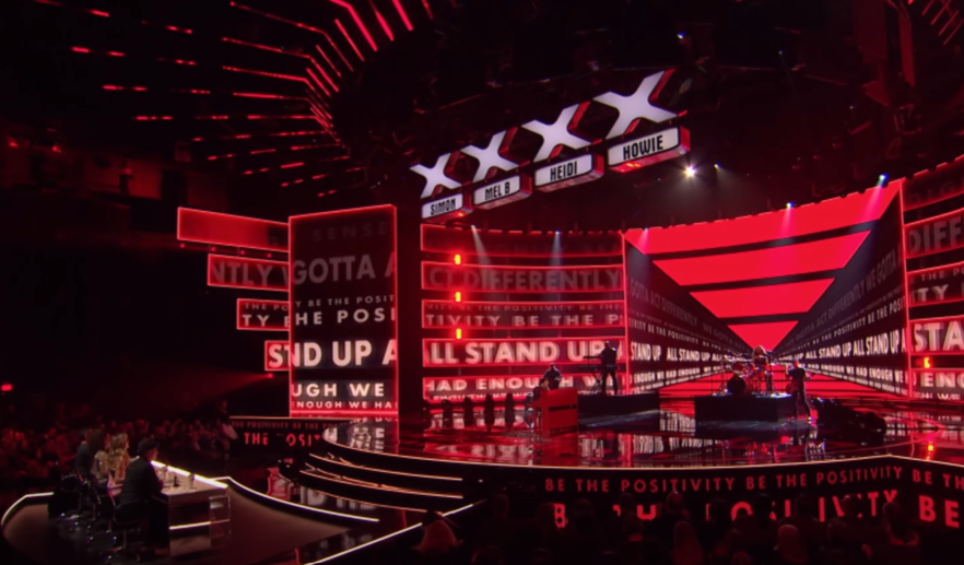 Full view of the "AGT" stage as Kodi Lee performs his original song "Change." The lighting is red and the screen displays messages like "Stand up" and "positivity"