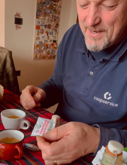 A man laughs as he holds a sugar packet in front of his lovebird. The bird is sitting on the dining table and is starting to open the pack of sugar with their beak.