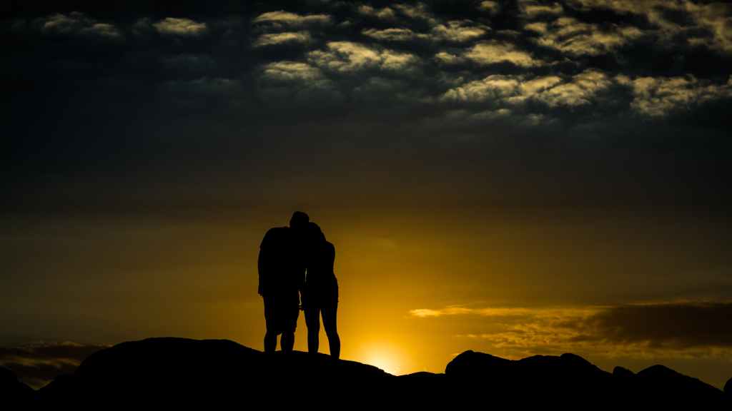 The shadow of a couple resting their heads on each other as they hold hands. The sun is setting, and it seems they're on a mountainside. 