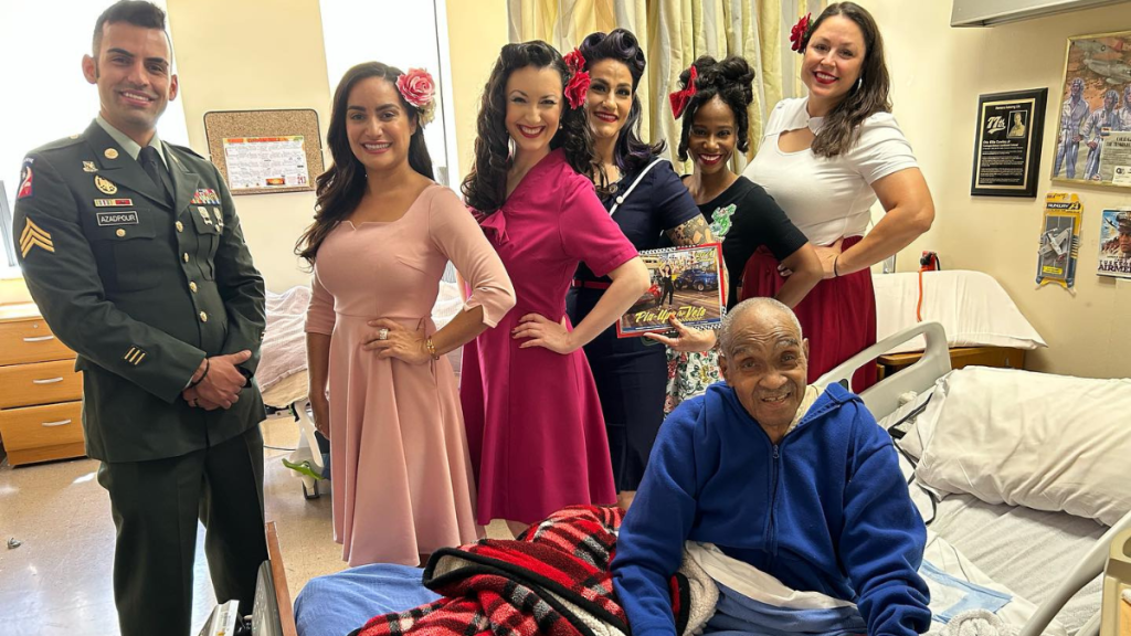 The Pin-Ups for Vets women smile and pose with a veteran in a hospital bed.